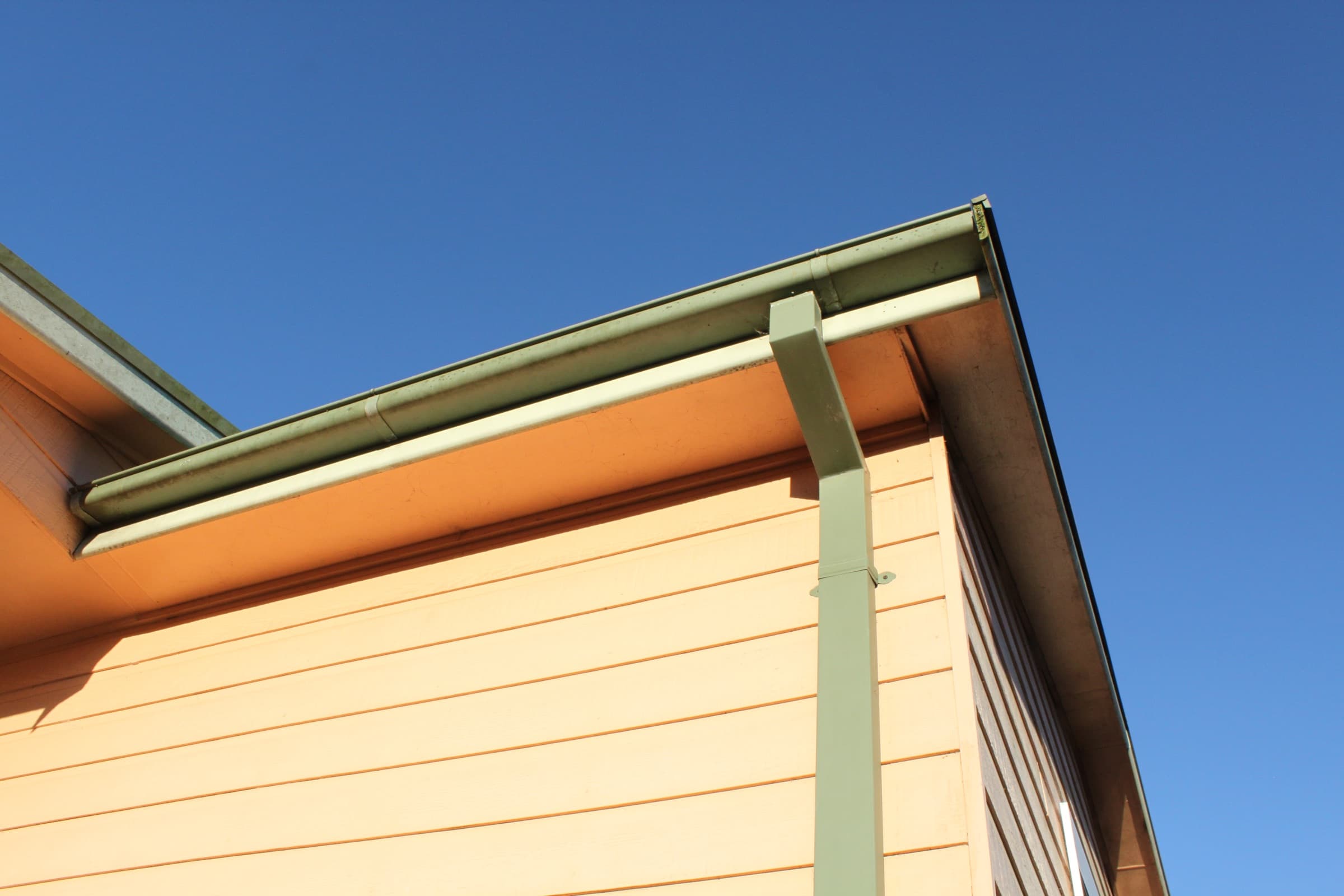 A close-up of a house roofline showing a rain gutter and downspout against a blue sky.